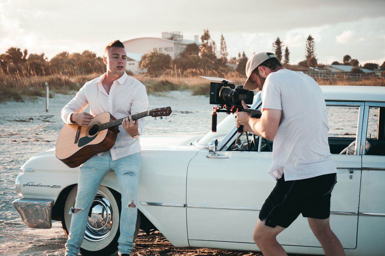 Musician plays guitar as a videographer films by a vintage car on a sunny beach